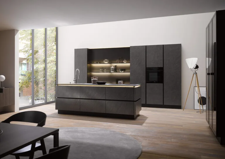 Isla de cocina en acabado gris pizarra con estantería abierta iluminada, columna de electrodomésticos y grandes ventanales